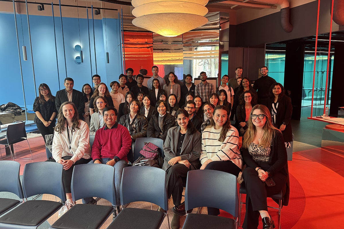 A diverse group of about 30 students in chairs and standing, gathered in a room for a meeting.