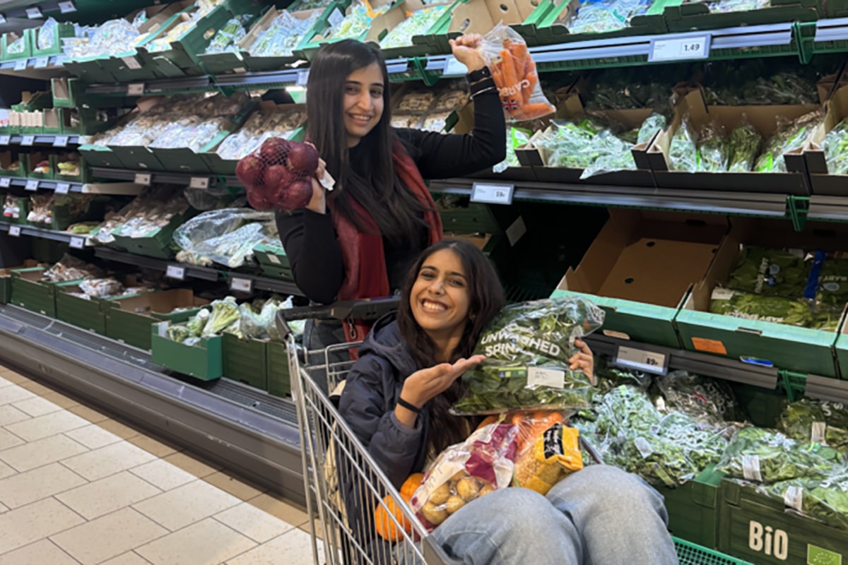 Pranshi is pushing a shopping trolley with her friend sitting in it down the vegetable aisle of a supermarket