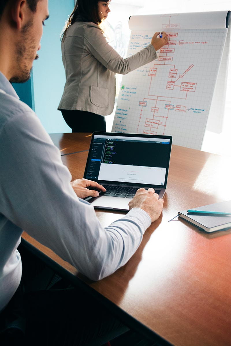 Person on laptop at desk with someone writing on a whiteboard in the distance
