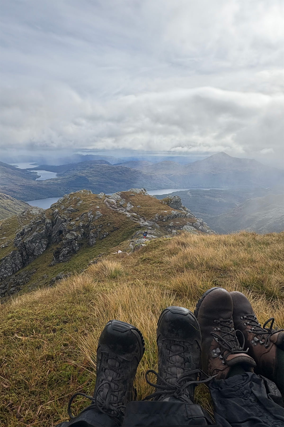 A moment of rest at the summit of Ben Vane. Two pairs of hiking boots are visible, overlooking a rugged landscape, with rocky peaks and a distant loch under a cloudy sky, conveying a sense of adventure.