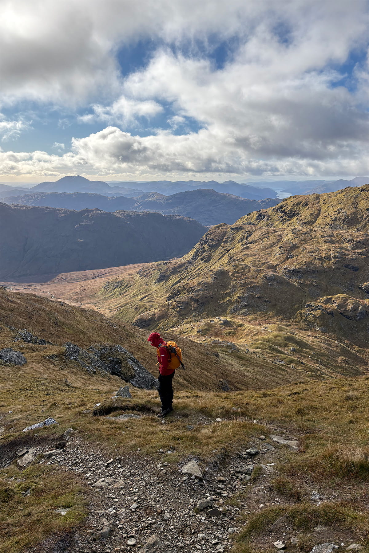 A person in a red jacket stands on a rocky mountain path overlooking Crianlarich. In the distance, a vast rugged landscape unfolds under a sky dotted with clouds and patches of blue.