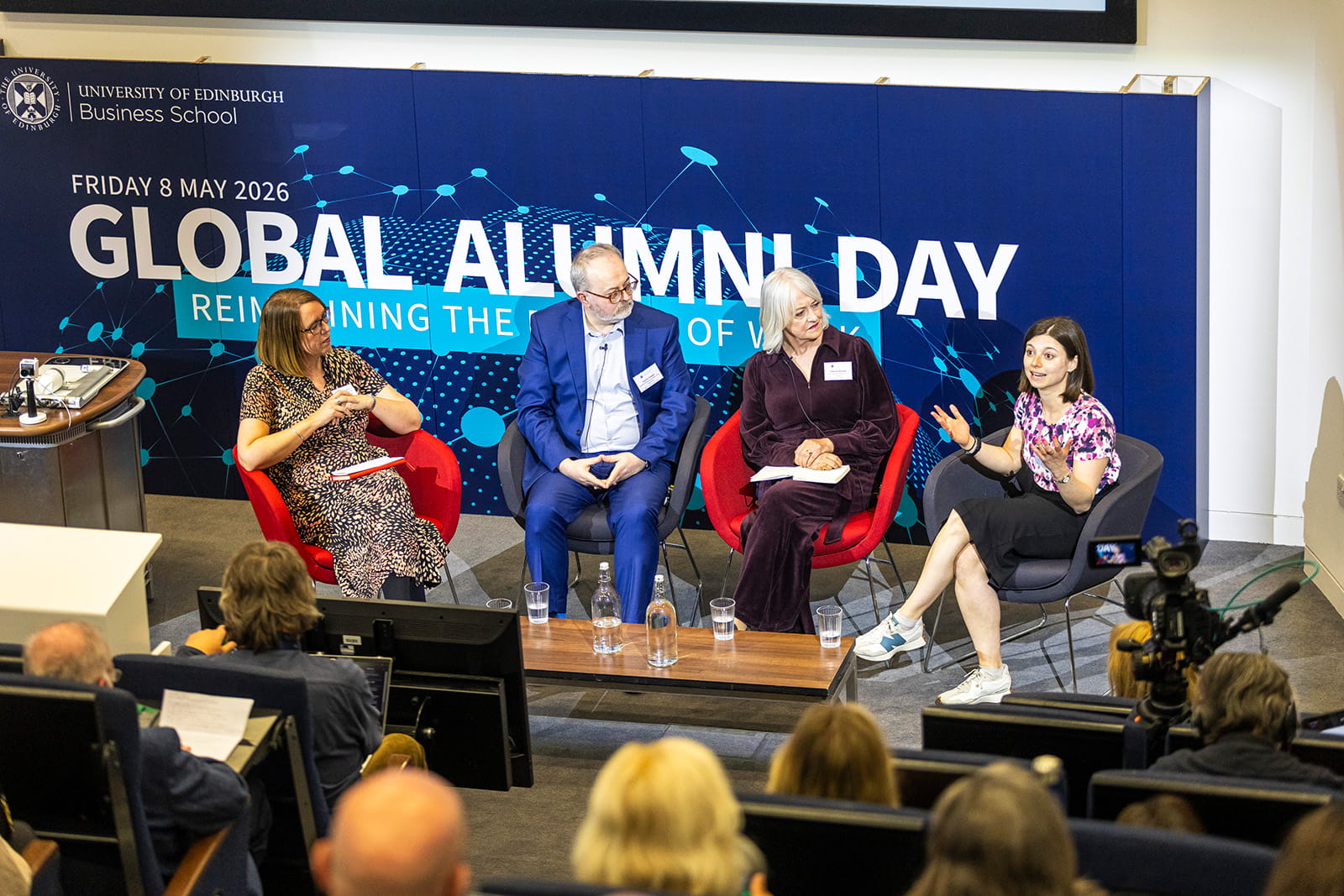 Four panelists sit on stage at Edinburgh Business School’s Global Alumni Day. The animated discussion involves diverse perspectives. The audience watches intently.