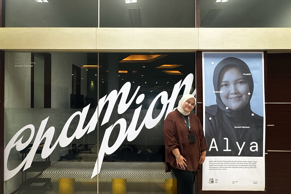 Student stands in front of a glass wall, beside a poster of themselves, with the word 