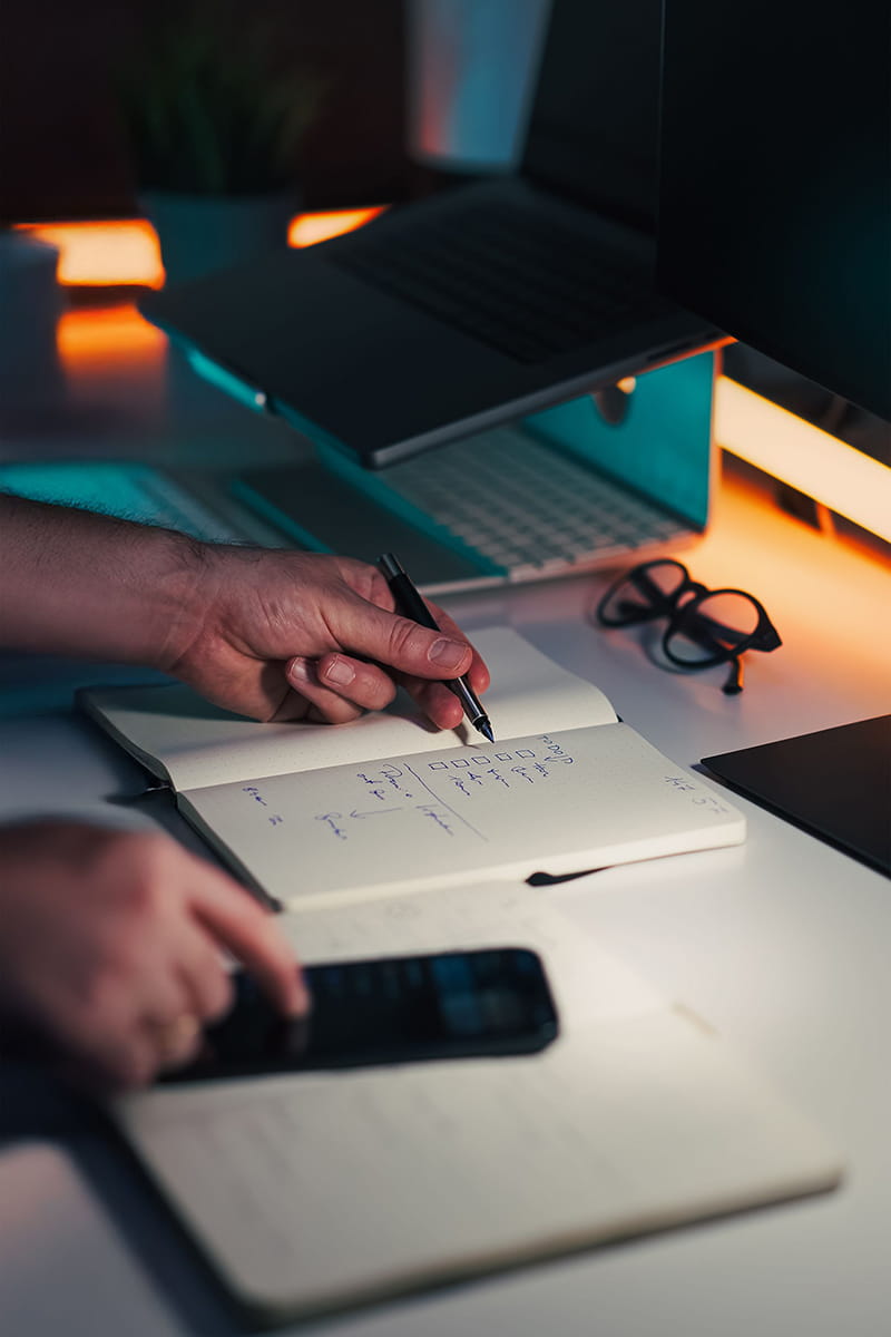 Person's hands writing in a notebook and using a mobile phone at a desk, with a computer and laptop present.