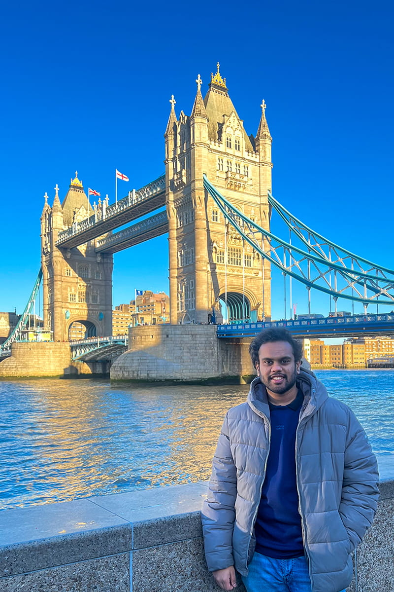Sidhanth standing in front of the Thames River with Tower Bridge visible in the background in London..