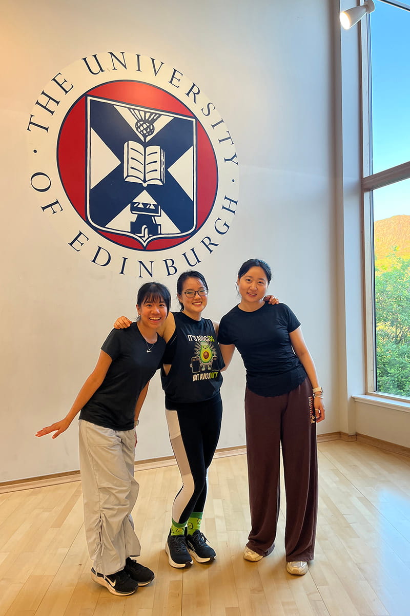 Three University of Edinburgh students pose together in front of a wall featuring the University of Edinburgh logo, located inside the University's gym