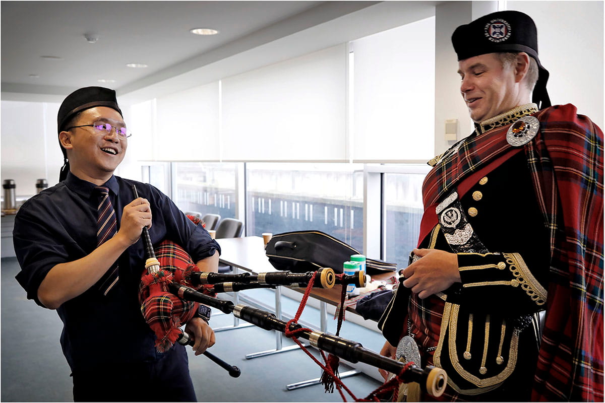  A bagpiper in full Highland dress standing next to a younger man in a suit who is holding the bagpipes