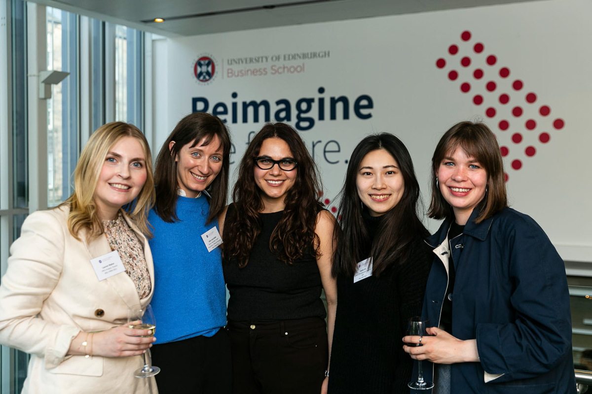 Five classmates stand together at Global Alumni Day, celebrating connections and reunions. They’re positioned in front of a backdrop with the text “Reimagine the Future”.
