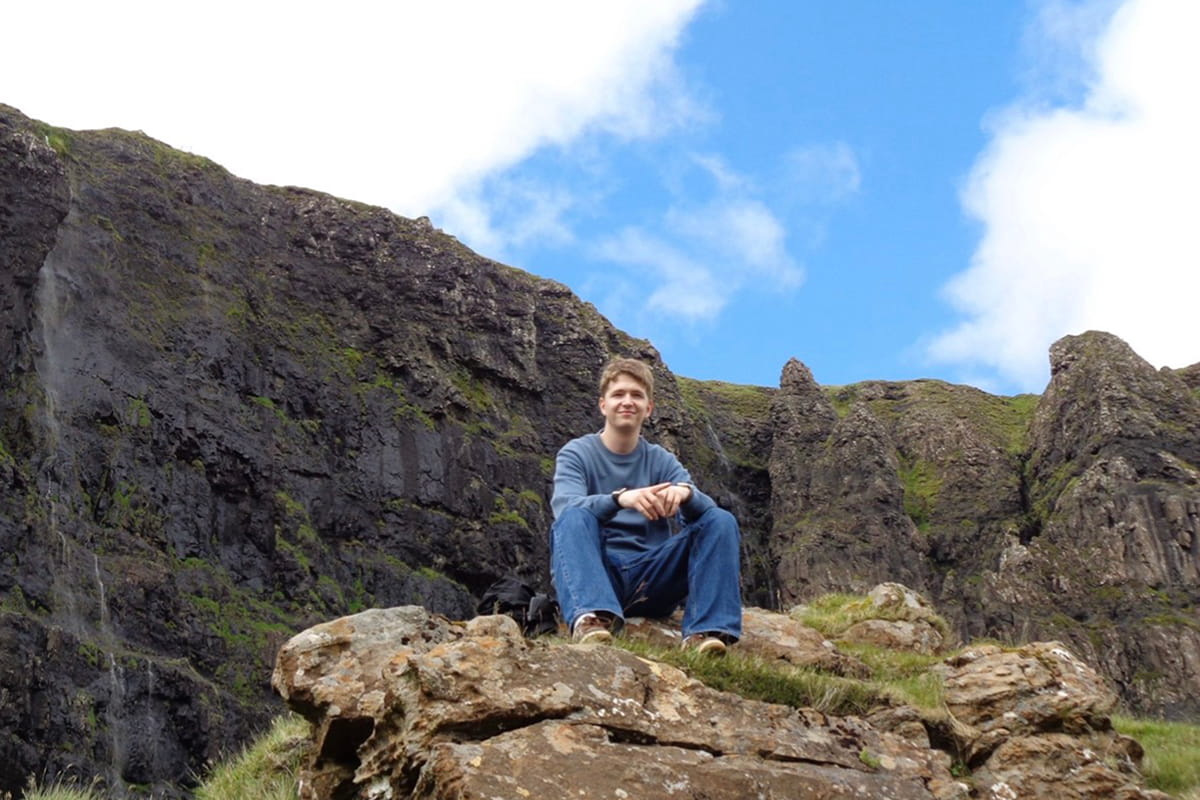 MSc student sitting on a rock with a dramatic background of cliffs behind him in Skye