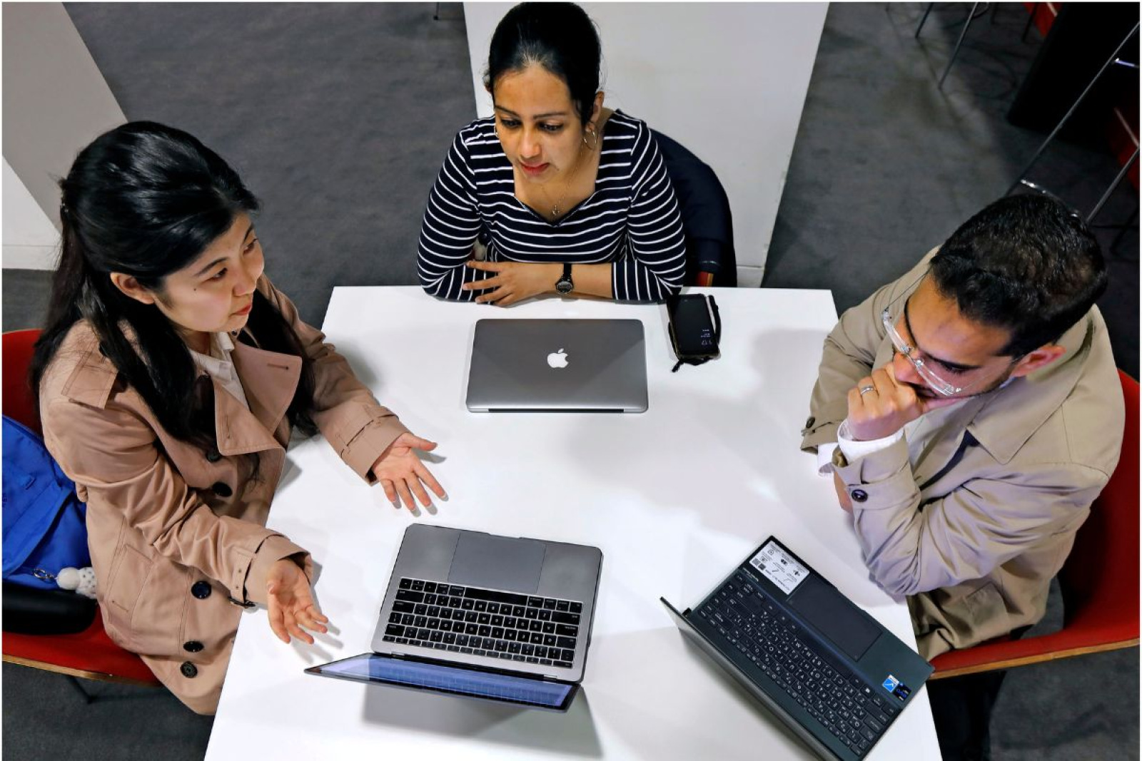 Three students seated at a desk looking at work on a laptop being presented by the student on the left.